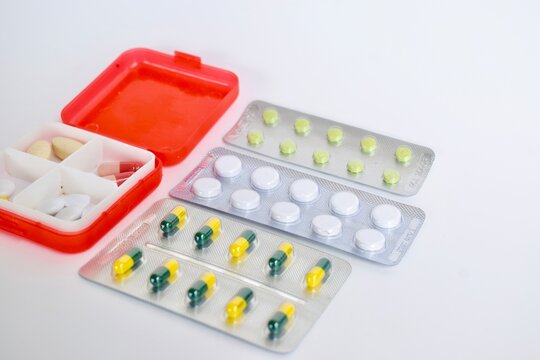 High angle close-up shot of an open red medical pill organizer next to assorted tablet and capsule blister packs on a white background.