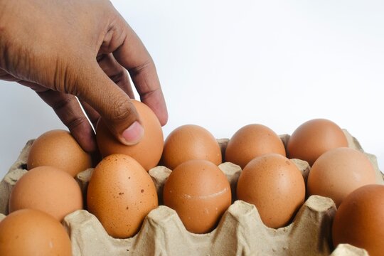 Close up of a man hand picking a fresh brown chicken egg from a cardboard carton box on a white background.