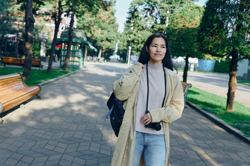 Naklejka premium Woman in light coat with camera walks through a sunny park, casual street style, greenery, and relaxed mood in an urban outdoor scene featuring bright light, breeze, and casual fashion vibe for