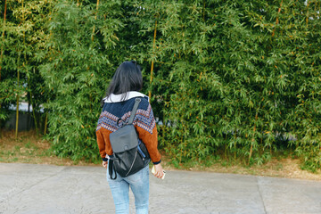 Naklejka premium Back view of a woman wearing a patterned knit sweater and backpack, strolling along a paved path beside a dense green hedge, conveying a casual outdoor commute and relaxed travel vibe