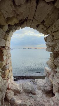 Vertical video: view through stone arch of Surlari Kalintilari fortress ruins toward Marmara Sea in Istanbul. Tanker ships visible on calm water under cloudy sky beyond ancient masonry frame.