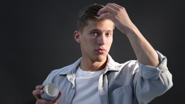 Handsome young man applying hair styling product onto his hair on dark grey background