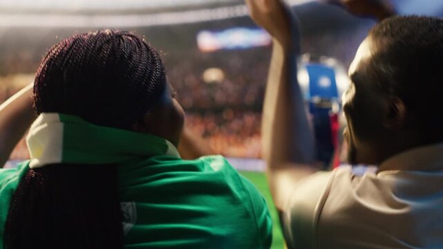 Football fans in stadium stands watching match unfolding on field, cheering for favorite team, showing soccer supporters looking at football pitch in packed arena crowd for fan culture content.