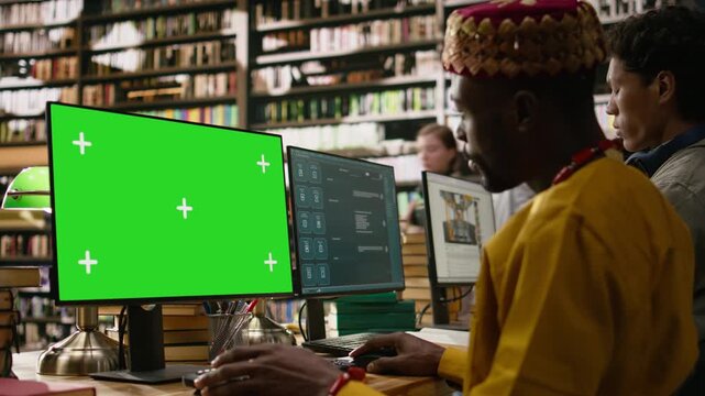 Green screen on desktop monitor near asian student typing at table, using mockup screen replacement on computer display, library workstation with books, for editable screen mockup and VFX use.