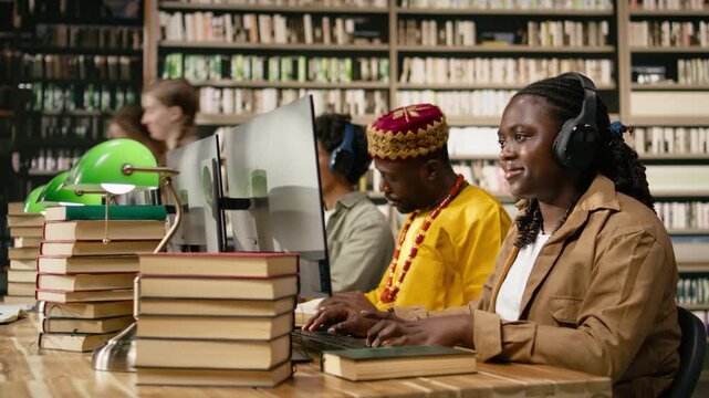 Black student wearing headphones reading open book at desktop on study desk while classmates study in library surrounded by shelves.