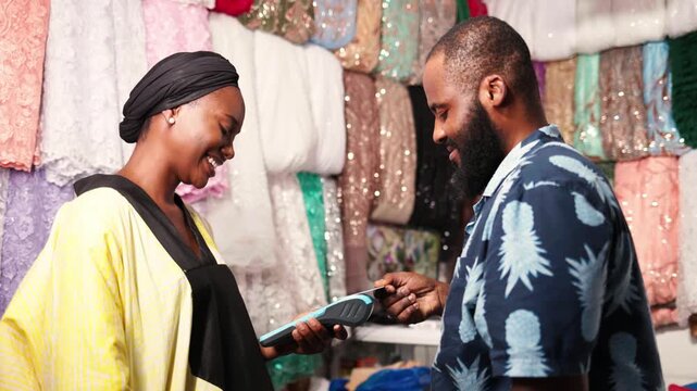 Smiling African businesswoman processing payment via pos terminal for customer in her fabric store