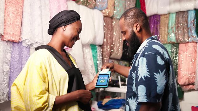 African woman small business owner smiling while customer pays using a pos terminal in her textile garment shop