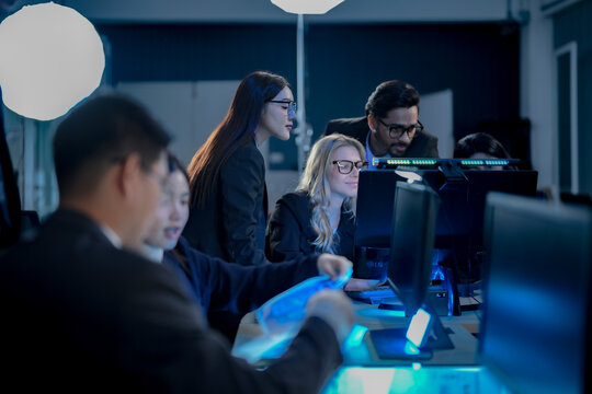Businesswoman leaning over a glowing desk to study documents with her blonde colleague underlining the key clause while a manager watches in a dim blue technology office as one team.