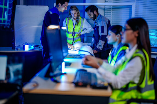 Engineers in high visibility vests at CAD workstations in a control room checking the level dimensions hard hats and radios on the desks around them as one team.