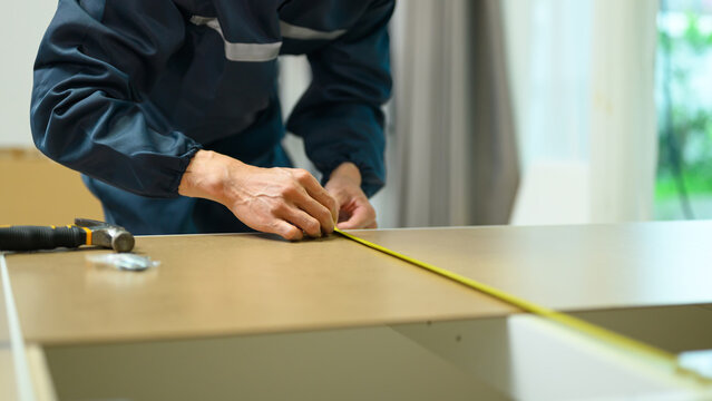 Worker in uniform measuring wooden board with tape measure during furniture assembly or home renovation