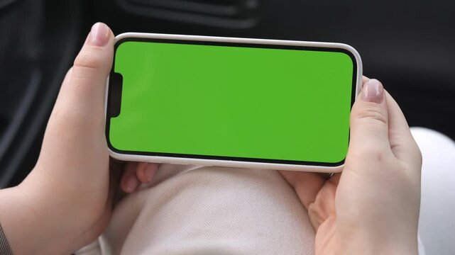 Hands hold phone with green screen, inside car. Fingers gently grip device, ready to interact. View shows partial lap and car interior. Ideal for app or video ads, mobile marketing, or UI design