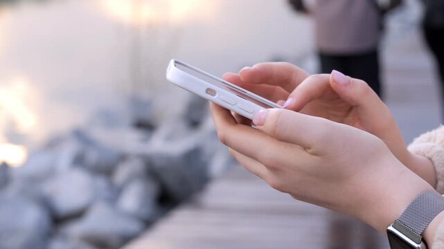 Close-up hands scroll smartphone near rocks. Person wears smartwatch, focused on screen. Outdoors with soft lighting, calm atmosphere. Ideal for social media, tech, or lifestyle content