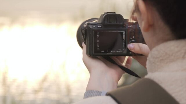 Person holds camera, focused on capturing scenic view. Warm light bathes scene, suggesting golden hour. Photographer wears cozy jacket, standing near water. Ideal for travel, nature.