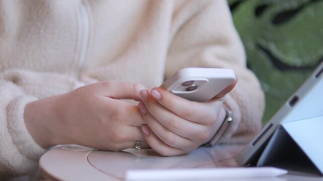 Close-up of hands holding a smartphone. Person wears cozy beige hoodie. Background shows laptop and green plant. Calm, focused mood suggests work or communication. Ideal for digital lifestyle