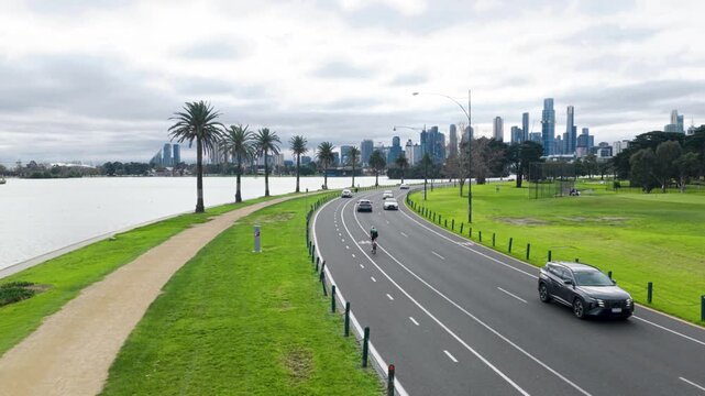 Aerial drone view along Albert Park Lake Road with cyclists and passing traffic beneath tree-lined streets, leading toward the Melbourne CBD skyline in the background.