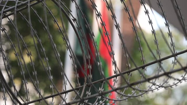 Coiled security razor wire stands in sharp focus as the BNP party flag hangs obscured in the soft background in Dhaka. Documents regional political conflicts, party crackdowns, and safety.