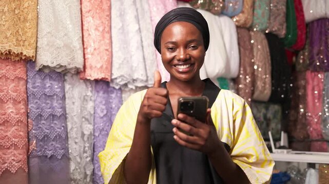 Smiling African woman entrepreneur showing thumbs up with smartphone in her thriving textile business