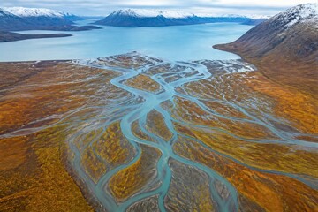 Naklejka premium Rivers connect with a big lake in a remote landscape. Mountains surround the water. The ground is a mix of green and brown colors. Clouds cover the sky.