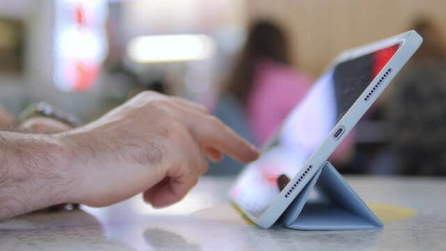 Hand gently taps a tablet screen. The device rests on a stand, angled for easy viewing. Background shows blurred cafe patrons, suggesting casual, everyday use