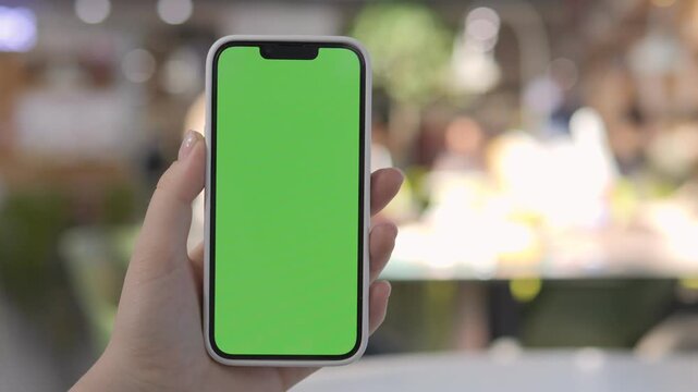 Hand holds smartphone with green screen. Background softly blurred, bokeh effect present. Person appears calm, focused on device. Ideal for app demos, digital marketing, or UI mockups
