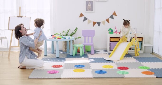 Asian mother kneeling on foam mat guiding preschool boy, preschool girl running across colorful stepping disc path near slide, slow motion active play, balance training, indoor daycare obstacle game