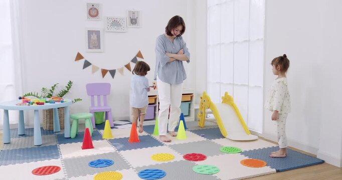 Asian female preschool teacher stands supervising obstacle course, preschool boy steps past cones, preschool girl pauses on foam mat, bright daycare playroom, balance pads, movement lesson, safe play