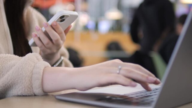 Person multitasking with laptop and phone. Hands typing on keyboard, phone held in other hand. Focus on work while staying connected. Suitable for remote work, digital communication