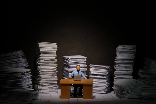 Miniature tiny people toy figure photography. Overload and work overtime concept. A businessman seat on a desk with pile of document, working until night at dark room with spotlight