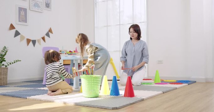 Asian female preschool teacher kneels coaching toddler boy, toddler girl leans stretching toss ring toward basket, cone markers on mat, bright daycare classroom, teamwork, active learning, gross motor
