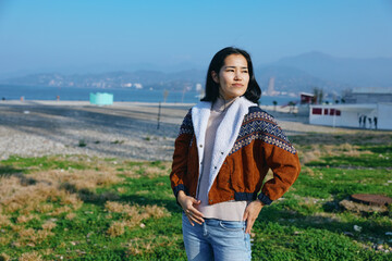 Fototapeta premium Woman standing outdoors in a burnt orange and white knit jacket with scarf, relaxed pose, exploring countryside scenery with meadow, hills and distant mountains under clear sky