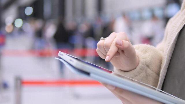 Person holds a tablet, using finger to touch screen. Background shows blurred crowd and barriers at event space. Focus remains on hand and device interaction
