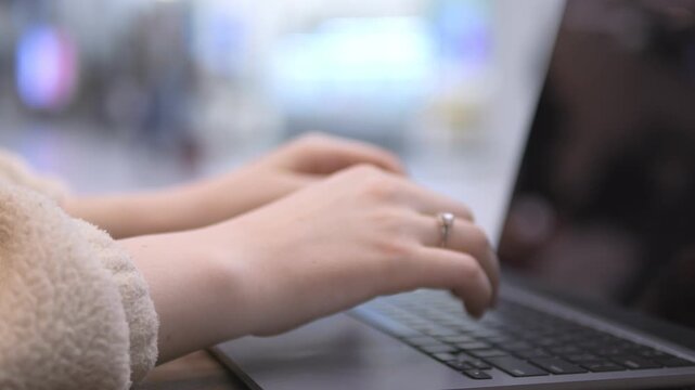 Hands type on laptop. Blurred background suggests public space. Woman wears ring. Soft lighting. No animals or plants visible