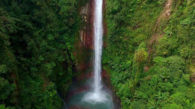 Cinematic drone aerial tilt-down and forward shot of a majestic high waterfall plunging into a deep turquoise canyon pool surrounded by lush tropical rainforest