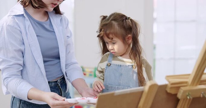 Adult asian mother prepares paint palette beside preschool daughter, guiding color choice for easel canvas, supportive parenting, creativity practice, fine motor skill exercise indoors, homeschool.