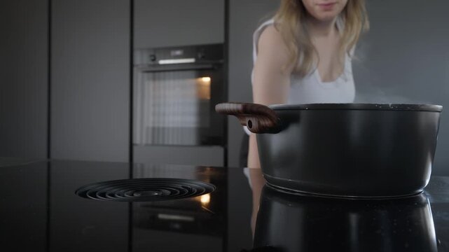 A woman prepares a meal in a sleek, dark minimalist kitchen. Steam rises from a black pot on an induction hob featuring an integrated downdraft vent. Authentic lifestyle scene.