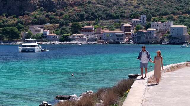 Couple walking hand in hand by the turquoise seashore, Yachting in Limeni village, traditional stone buildings and mountains in the background, Static shot