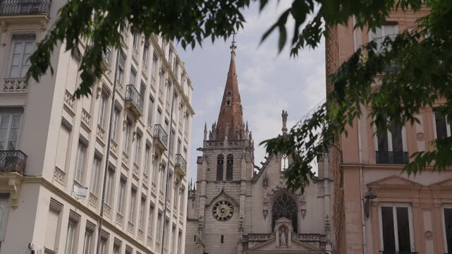 A cinematic shot of a historic gothic church steeple in Lyon, France, framed by traditional European apartment buildings and green summer leaves.