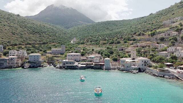 Drone circles around sun drenched Limeni village on crystal clear turquoise waters with stone houses and steep lush green mountains in the backdrop under cloudy sky, Southern Peloponnese