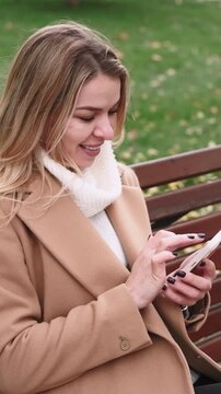 Vertical video. Sending message. Urban nature. Smiling female scrolling online social media chat on mobile telephone relaxing on park bench.