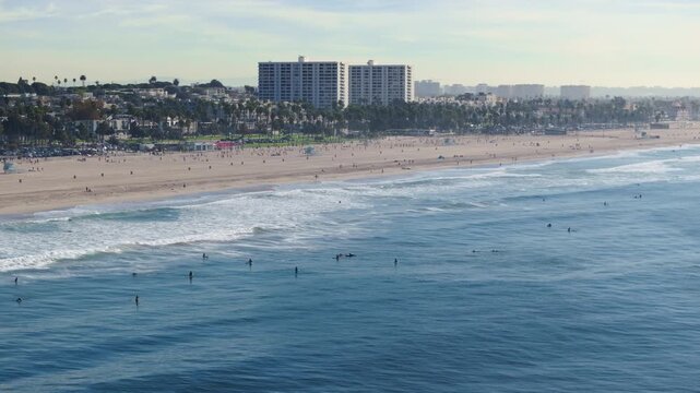 Aerial view of Santa Monica California USA coastline featuring surfers waiting for waves in the Pacific Ocean, sandy beach with beachgoers, palm trees, high-rise buildings, and hazy city skyline.