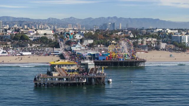 Santa Monica, California, USA - JANUARY 5, 2025: Aerial view of Santa Monica Pier with colorful ferris wheel and roller coaster rides extending over Pacific Ocean with beach and mountain backdrop.