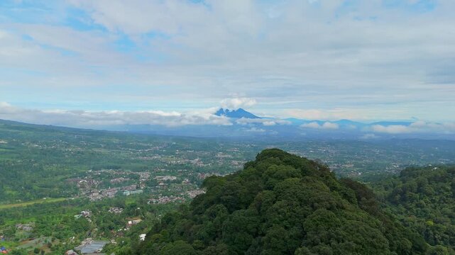 Cinematic drone aerial wide shot of majestic mountain peak with rolling clouds overlooking a lush green valley and rural town, Indonesia