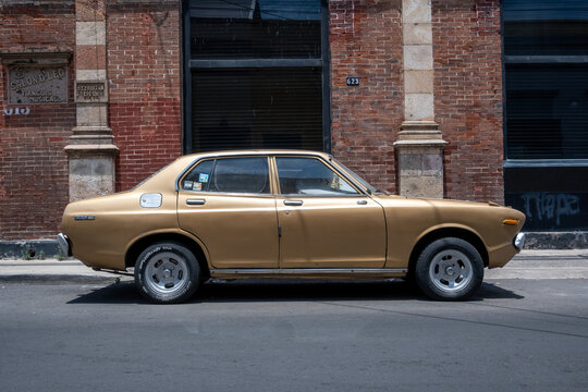 Guadalajara, Mexico - May 9 2026: A vintage gold sedan, possibly a Datsun, parked on a sunlit street in Guadalajara, Mexico. Old brick building and classic architecture