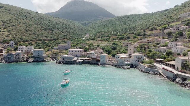 Drone shot of sun drenched Limeni village on crystal clear turquoise waters with stone houses and steep lush green mountains in the backdrop under cloudy sky, Southern Peloponnese