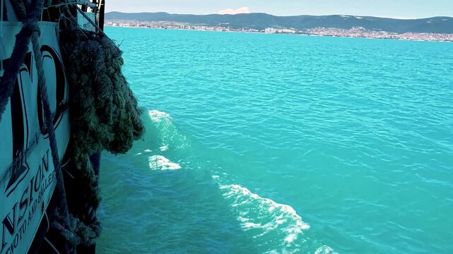 A side view from a moving boat captures vibrant turquoise sea water creating a gentle white wake, with a textured rope hanging in the foreground.
