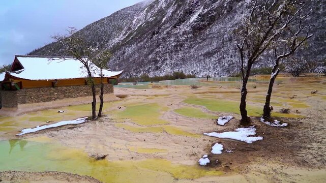 A wide slow panning shot of golden-yellow mineral terraces and light green pools next to a traditional stone-walled pavilion below snow-dusted slopes.