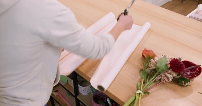 Florist working. Wrapping bouquet. Unrecognizable professional female using scissors cutting paper for dahlia flowers on wooden table.