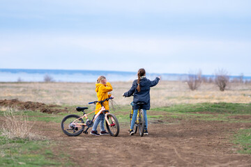 Fototapeta premium Two happy sisters or girlfriends on a cloudy spring or autumn day ride bicycles in nature along a dirt road against the backdrop of the sea or lake