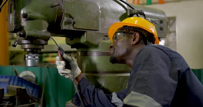African adult male technician in hardhat inspects milling machine close up, holding air blow gun near spindle during maintenance cleaning and safety check in workshop slow motion scene