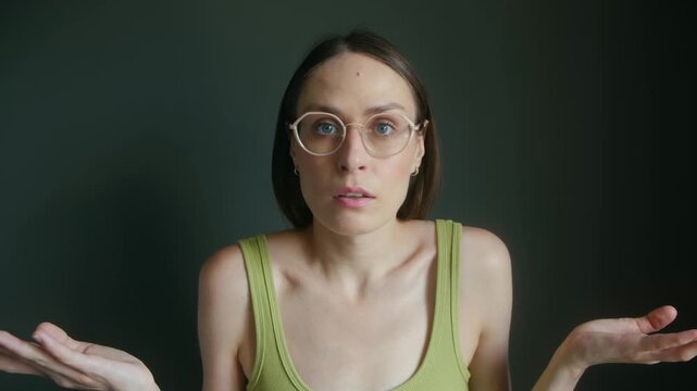 Puzzled young woman with eyeglasses shrugging shoulders making clueless gesture, expressing confusion, doubt, uncertainty while looking at camera. Confused woman with glasses shrugging shoulders